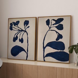 May include: Two rectangular framed wall art prints featuring dark blue abstract botanical silhouettes against a textured off-white background. The artwork displays thick, raised paint strokes forming organic plant shapes with stems and leaves.