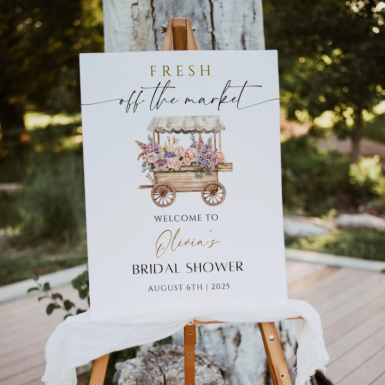 May include: A white sign with gold lettering that says "Fresh off the market" and features a watercolour illustration of a flower cart. The sign also says "Welcome to Olivia's Bridal Shower August 6th | 2025".
