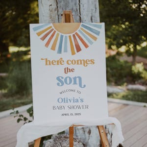 May include: A white welcome sign with a watercolor sun design in shades of orange, yellow, and blue. The text reads "Here comes the son" with a blue and white script font. Below the text is "Welcome to Olivia's Baby Shower" and the date "April 15, 2025".