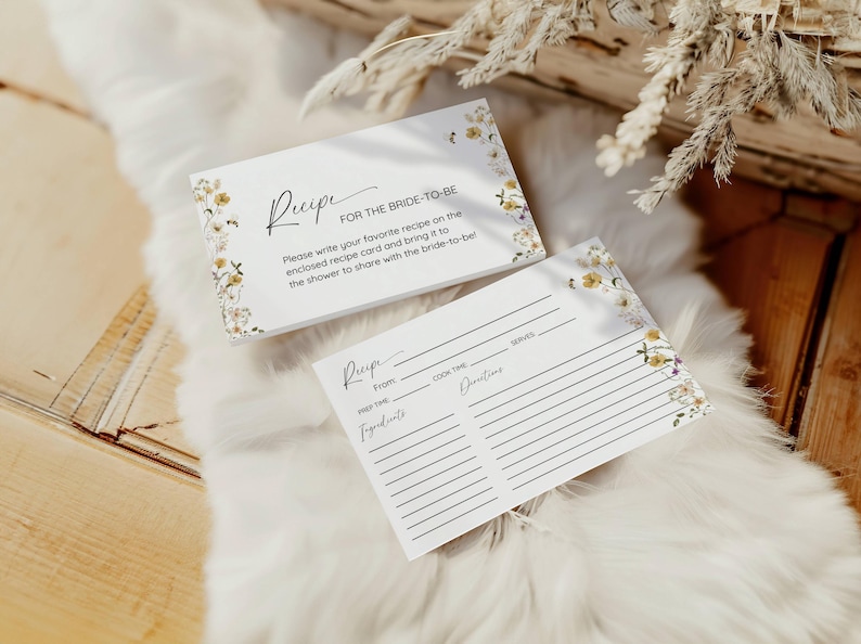 Puede incluir: Dos tarjetas de recetas blancas con dise&ntilde;os florales. La tarjeta superior tiene el texto "Receta para la novia" e instrucciones para escribir una receta favorita. La tarjeta inferior es una plantilla de receta en blanco con secciones para "De", "Tiempo de preparaci&oacute;n", "Tiempo de cocci&oacute;n", "Porciones", "Ingredientes" y "Instrucciones".