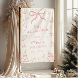 May include: A baby shower banner with a light background and pink illustrations of snowflakes, a bow, and winter-themed items. The text reads "Baby it's cold outside" and welcomes guests to Olivia's baby shower on August 26th, 2025.