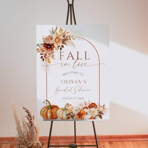 以下が含まれることがあります： 黒いイーゼルに立てられた白い看板。テキストは「FALL in love」と「Welcome to Olivia's Bridal Shower August 6th, 2025」です。秋をテーマにしたフラワーアレンジメントとカボチャで飾られています。