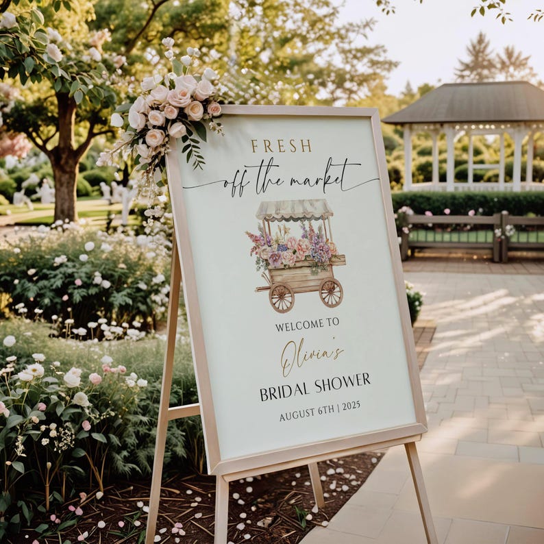 May include: A white sign with a wooden frame on an easel. The sign has a floral illustration of a flower cart with the text "FRESH off the market" in gold script. The text "Welcome to Olivia's Bridal Shower August 6th | 2025" is printed below the illustration.
