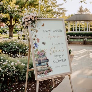 Pode incluir: Um cavalete de madeira segura uma placa com detalhes florais e borboletas. A placa diz "A New Chapter begins soon" e d&aacute; as boas-vindas a "Olivia's Bridal Shower" em 26 de agosto de 2025. Um buqu&ecirc; de rosas adorna o topo.
