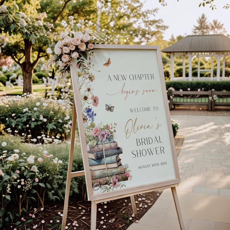 Pode incluir: Um cavalete de madeira segura uma placa com detalhes florais e borboletas. A placa diz "A New Chapter begins soon" e d&aacute; as boas-vindas a "Olivia's Bridal Shower" em 26 de agosto de 2025. Um buqu&ecirc; de rosas adorna o topo.