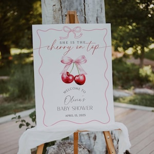 May include: A white sign with pink and red accents announcing a baby shower. The sign reads "She is the cherry on top. Welcome to Olivia's Baby Shower. April 15, 2025".