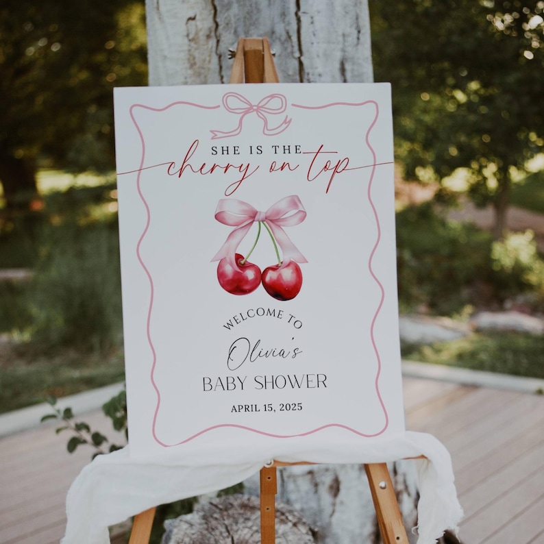May include: A white sign with pink and red accents announcing a baby shower. The sign reads "She is the cherry on top. Welcome to Olivia's Baby Shower. April 15, 2025".