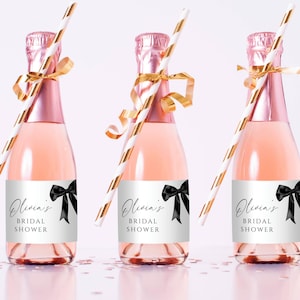 May include: Three pink champagne bottles with gold ribbons and striped straws. Each bottle has a label that reads "Olivia's Bridal Shower" with a black bow graphic. The bottles are arranged on a light pink surface.