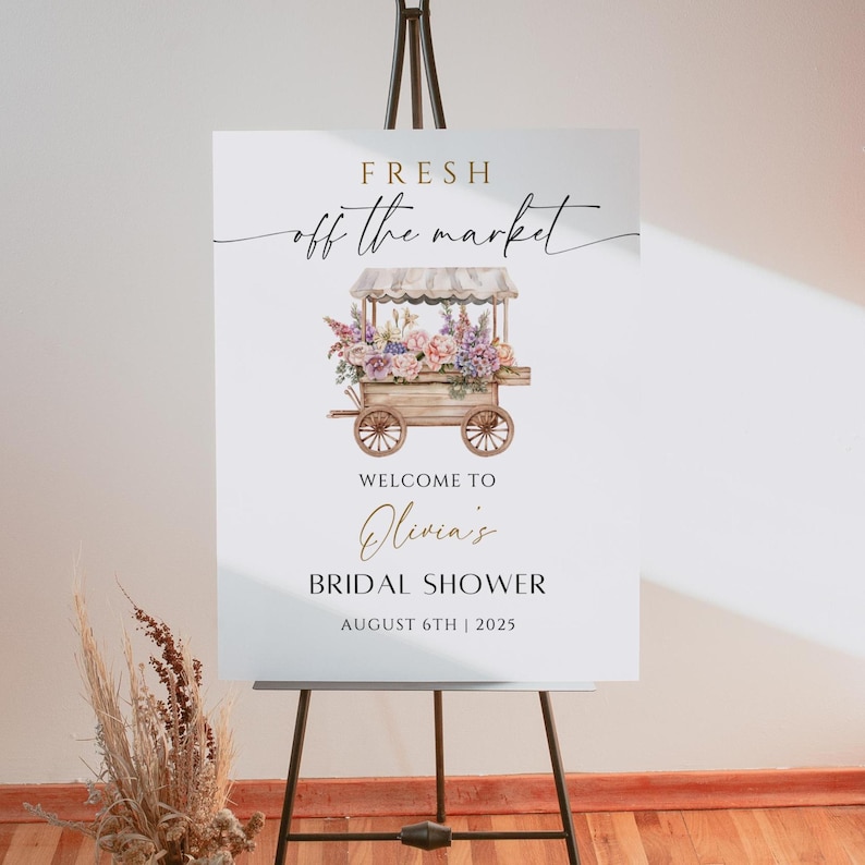May include: A white sign with gold lettering announcing a bridal shower. The sign features a watercolour illustration of a flower cart with a banner that reads "Fresh off the market". The text on the sign reads "Welcome to Olivia's Bridal Shower August 6th | 2025".