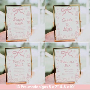 May include: Four framed signs with gold borders and pink text. The signs read "Diaper Raffle", "Cards & Gifts", "Pacifier Hunt", and "Mom-osa Bar". Each sign has a pink bow and winter-themed illustrations. The signs are 5 x 7 inches and 8 x 10 inches.