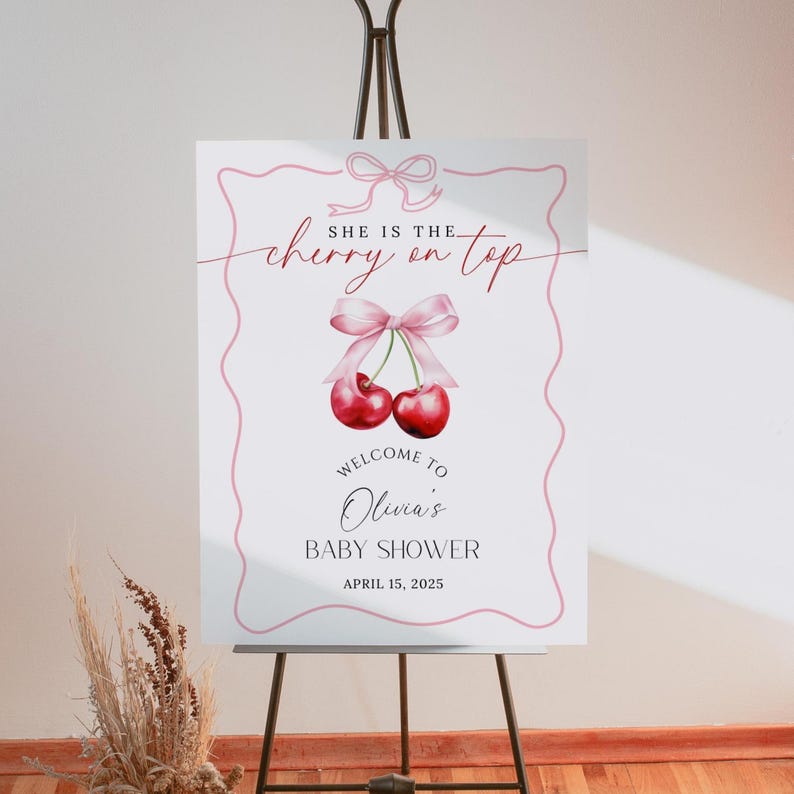 May include: A white baby shower sign with pink and red cherries and a pink ribbon. The sign says "She is the cherry on top" and "Welcome to Olivia's Baby Shower April 15, 2025".