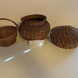 May include: Three brown woven baskets, one with a handle, one with a lid, and one with a rounded shape. The baskets are made of natural materials and have a rustic appearance.