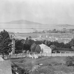 May include: A black and white photograph of a large house with a greenhouse in the foreground. The house is surrounded by trees and bushes. The photo was taken from a higher vantage point, and the background shows a body of water and a distant city.