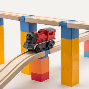 May include: A red and black toy steam locomotive engine sits on a wooden train track elevated by a series of interlocking plastic support blocks in yellow, blue, orange, and red. The track system features natural wood rails and blue plastic connectors.
