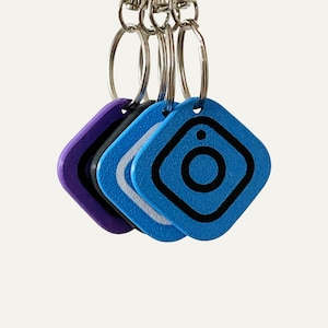 May include: A collection of keychains in various colors, including purple, black, and blue. Each keychain features a square design with a rounded edge and a black camera icon. The keychains are attached to silver key rings.
