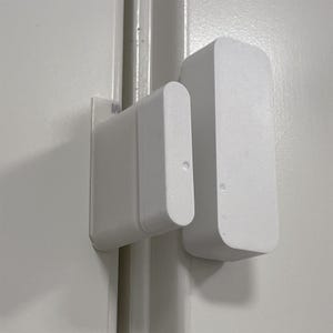 May include: A white door sensor with two parts, one mounted on the door frame and the other on the door. The sensor is used to detect when a door is opened or closed.