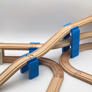 May include: Wooden train track set with blue supports. The track is arranged in a curved and elevated configuration, creating a bridge-like structure. The supports are labeled "WOODCRAFT WORKSHOP".