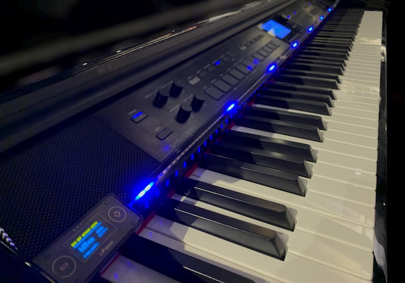 Peut inclure: Un clavier num&eacute;rique noir dot&eacute; d'une s&eacute;rie de touches blanches et noires, d'un panneau de commande avec divers boutons et molettes, et d'un petit appareil rectangulaire fix&eacute; sur le c&ocirc;t&eacute; avec un &eacute;cran num&eacute;rique bleu et le texte L-Piano.