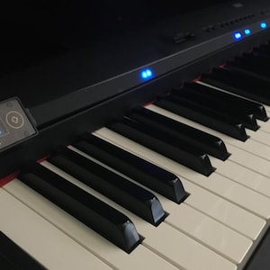 May include: Close-up of a black digital piano with white and black keys. The piano has a small screen displaying text and blue lights along the top. The text on the screen reads "i-Piano".