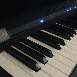 Op de afbeelding: Een zwart digitaal pianoklavier met witte en zwarte toetsen, een rij oplichtende blauwe indicatielampjes op het bovenpaneel en een klein rechthoekig bedieningsscherm aan de linkerkant met de tekst i-Piano en zichtbare menu-instellingen.
