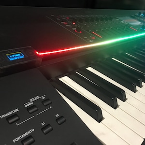 Peut inclure: Un clavier &eacute;lectronique noir dot&eacute; de touches blanches et noires, d'un panneau de contr&ocirc;le avec des boutons marqu&eacute;s Octave, Transpose et Portamento, et d'un petit &eacute;cran num&eacute;rique affichant le texte I-PIANO avec une bande lumineuse rouge et verte.