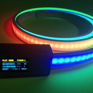 Peut inclure: Une bande lumineuse LED flexible enroul&eacute;e en cercle, &eacute;mettant une lumi&egrave;re vive verte, jaune et bleue. Un bo&icirc;tier de commande rectangulaire noir est attach&eacute;, avec un &eacute;cran num&eacute;rique affichant le texte : PLAY MODE PIANO-1, BRIGHTNESS (+) 31, COLORSCHEME (-) 1, RESET (+) & (-) 10sec.