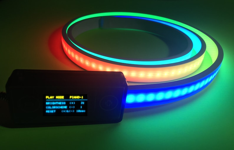 Peut inclure: Une bande lumineuse LED flexible enroul&eacute;e en cercle, &eacute;mettant une lumi&egrave;re vive verte, jaune et bleue. Un bo&icirc;tier de commande rectangulaire noir est attach&eacute;, avec un &eacute;cran num&eacute;rique affichant le texte : PLAY MODE PIANO-1, BRIGHTNESS (+) 31, COLORSCHEME (-) 1, RESET (+) & (-) 10sec.