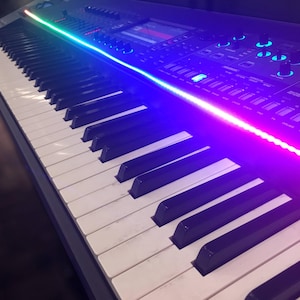 Peut inclure: Un clavier synth&eacute;tiseur de musique &eacute;lectronique noir dot&eacute; d'une longue bande lumineuse LED horizontale passant du vert au bleu au magenta. Le panneau de commande comprend un &eacute;cran d'affichage num&eacute;rique central, plusieurs boutons rotatifs et poussoirs.