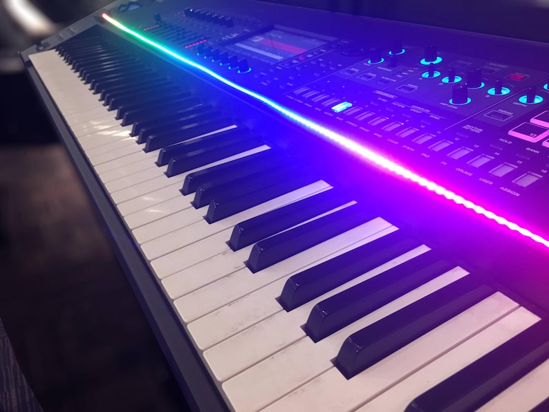 Peut inclure: Un clavier synth&eacute;tiseur de musique &eacute;lectronique noir dot&eacute; d'une longue bande lumineuse LED horizontale passant du vert au bleu au magenta. Le panneau de commande comprend un &eacute;cran d'affichage num&eacute;rique central, plusieurs boutons rotatifs et poussoirs.