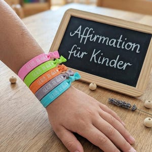 Könnte beinhalten: Fünf elastische Haarbänder in Pink, Limettengrün, Orange, Lavendel und Türkis, jeweils mit dem weißen Schriftzug "GLAUB AN dich" und einem kleinen Herzsymbol. Die Bänder werden um ein Handgelenk getragen, neben einer kleinen Kreidetafel.