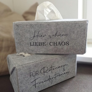 Könnte beinhalten: Zwei rechteckige Taschentuchboxen aus hellgrauem Filz, die vertikal gestapelt sind. Die obere Box trägt den schwarzen Schriftzug "Hier wohnen LIEBE & CHAOS" und die untere den schwarzen Schriftzug "FÜR Rotznasen & Freudentränen" mit weißen Tüchern.