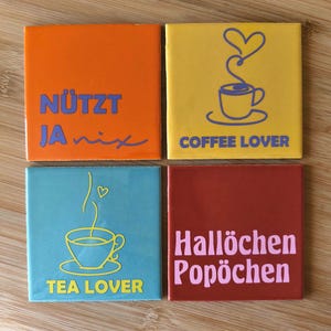 Könnte beinhalten: Ein Set aus vier quadratischen Untersetzern in Orange, Gelb, Türkis und Kastanienbraun. Der orangefarbene Untersetzer trägt die Aufschrift NÜTZT JA nix. Der gelbe zeigt eine Kaffeetasse mit Herz und den Text COFFEE LOVER. Der türkise zeigt eine Teetasse und TEA LOVER. Der kastanienbraune trägt die Aufschrift Hallöchen Popöchen.