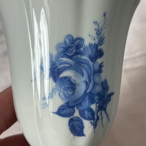 Puede incluir: Taza de té de porcelana blanca con un diseño floral azul. La taza tiene un borde acampanado y una base de pedestal. El patrón floral azul incluye rosas y otras flores. La taza de té está sostenida por una mano.