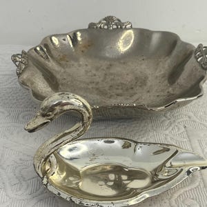 Puede incluir: Un plato de metal plateado con forma de cisne con un cuello curvo y un cuenco de metal plateado separado con bordes festoneados y asas decorativas. Ambos artículos muestran signos de desgaste y deslustre en sus superficies metálicas.