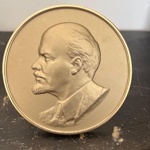 May include: A gold-coloured, circular medallion depicting a man's profile, facing left, with a beard and detailed facial features. The medallion has a raised border. The man's hair and facial features are finely detailed.