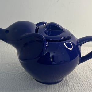 Può includere: Una teiera in ceramica blu scuro lucido a forma di elefante. La teiera presenta un beccuccio a forma di proboscide, ampi pannelli laterali a forma di orecchie e un coperchio rimovibile con una piccola maniglia. Il corpo è arrotondato con un manico.