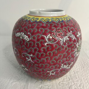 Peut inclure: Un vase rond en céramique rouge avec un bord blanc et une bande jaune. Le vase est décoré d'un motif répétitif de motifs floraux blancs et de volutes. Le vase est un objet décoratif.