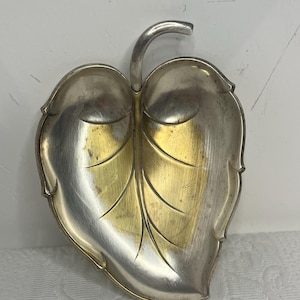Può includere: Un piatto a forma di foglia color argento con un centro dorato. Il piatto ha un gambo curvo e venature delle foglie dettagliate. Il piatto in metallo viene probabilmente utilizzato per scopi decorativi o come piccolo vassoio da portata.