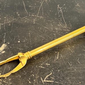 Op de afbeelding: Een goudkleurige metalen suikertang met een lange, slanke steel, een ronde drukknop aan het uiteinde en een veermechanisme dat twee gebogen, bloembladachtige grijpers bedient. Het gereedschap heeft een gladde afwerking met subtiele lijngravures.