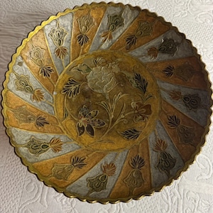Op de afbeelding: Een ronde decoratieve metalen schaal met een geschulpte rand en een centraal medaillon met een gegraveerd bloemmotief. Het oppervlak is verdeeld in afwisselende goud- en zilverkleurige panelen, elk versierd met een herhalend bloemmotief.
