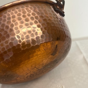 May include: A copper-colored, hammered metal pot with a rounded body and a small handle. The pot has a textured surface with a pattern of small, evenly spaced indentations. The pot is likely vintage or antique.