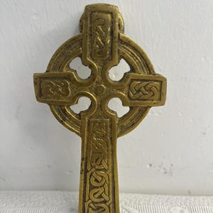 Könnte beinhalten: Ein keltisches Kreuz aus Messing mit komplizierten, geprägten Knotenmustern auf den vertikalen und horizontalen Armen. Das Design umfasst einen zentralen kreisförmigen Ring, der die vier Arme verbindet, mit einer texturierten, gealterten Metalloberfläche.