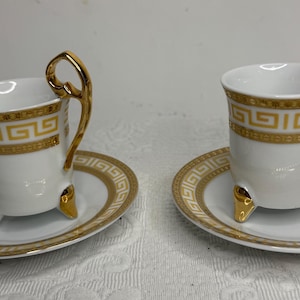 Może przedstawiać: Dwie białe filiżanki do espresso z porcelany wraz z pasującymi spodkami, ozdobione złotym wzorem greckiego klucza na krawędziach. Każda filiżanka posiada trzy małe złote nóżki oraz charakterystycznie wyprofilowane złote ucho.