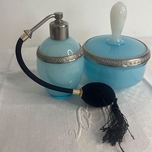 Könnte beinhalten: Ein Vintage-Kosmetikset mit einer hellblauen Parfümflasche mit einem schwarzen Zerstäuber und einem passenden Glas mit Deckel. Beide Teile haben silberfarbene Verzierungen. Der Zerstäuber hat einen schwarzen Ball und eine Quaste.
