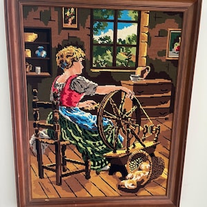 May include: A framed tapestry depicts a woman using a spinning wheel. The artwork features a woman in a red vest and blue skirt, seated in a wooden chair. The scene includes a window with a view of trees and a basket of bread.