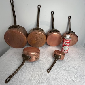 May include: A collection of copper saucepans with long, dark handles. The pans vary in size, with a small can of copper cleaner visible. The copper has a warm, reddish-brown hue, suggesting age and use. The handles have a loop for hanging.
