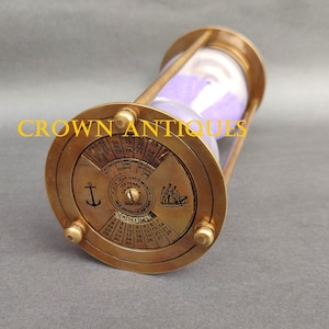 May include: Brass hourglass with a perpetual calendar on the base. The hourglass has a purple sand and a clear glass bulb. The calendar is a rotating dial with the months of the year and days of the week.