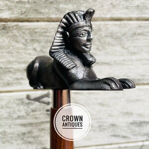 May include: A wooden walking stick with a black metal topper featuring a stylized sphinx head. The topper is detailed with a crown and a pharaoh's headdress. The stick has a silver band and the words "CROWN ANTIQUES" are printed on a white circle.
