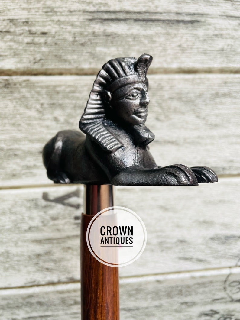 May include: A wooden walking stick with a black metal topper featuring a stylized sphinx head. The topper is detailed with a crown and a pharaoh's headdress. The stick has a silver band and the words "CROWN ANTIQUES" are printed on a white circle.