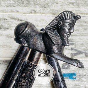 May include: A black walking stick with a detailed carved design and a decorative metal head shaped like a sphinx. The head is detailed with a pharaoh's headdress and facial features. The text "CROWN ANTIQUES" is visible on the stick.